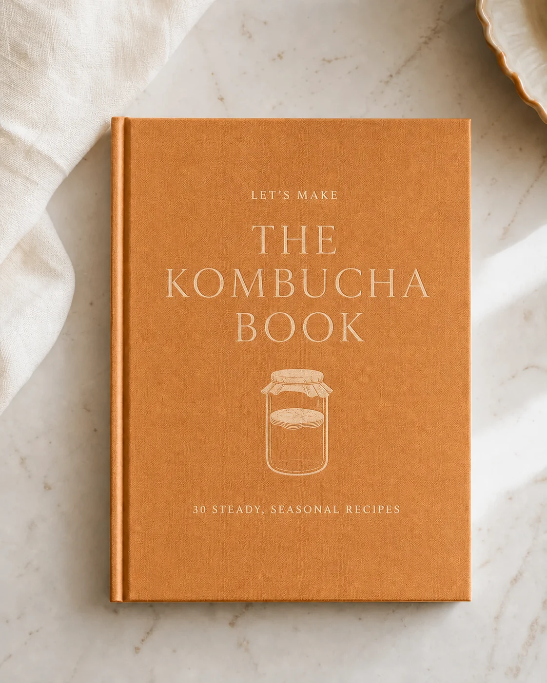 The Kombucha Book shown as a warm amber hardcover on a pale farmhouse kitchen counter